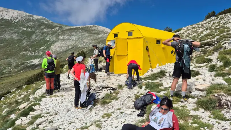 Gruppo di escursionisti vicino a un giallo blockhaus Majella, su un sentiero roccioso con montagne verdi sullo sfondo.